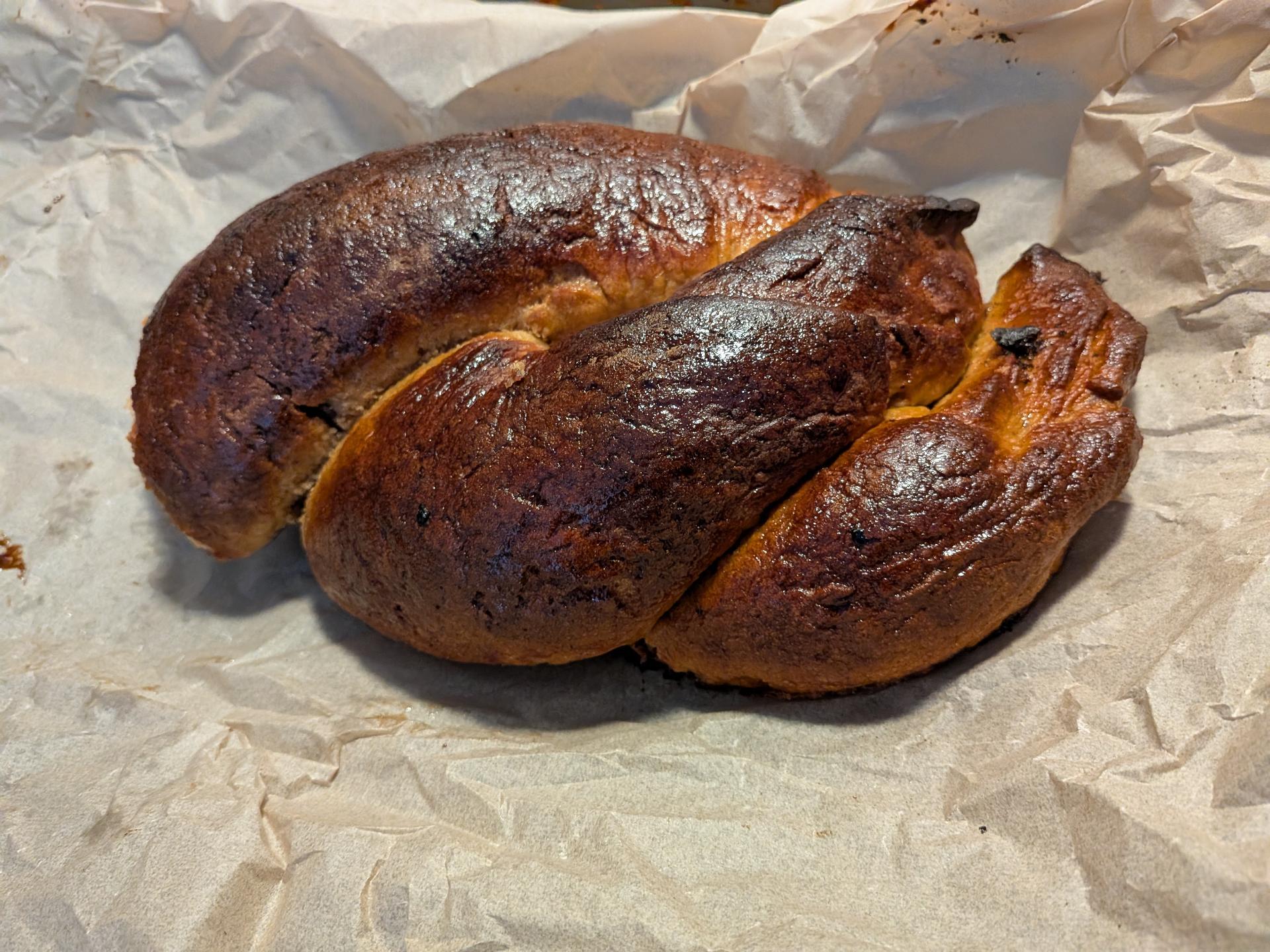 A dark, poorly braided loaf of challah bread on parchment paper.