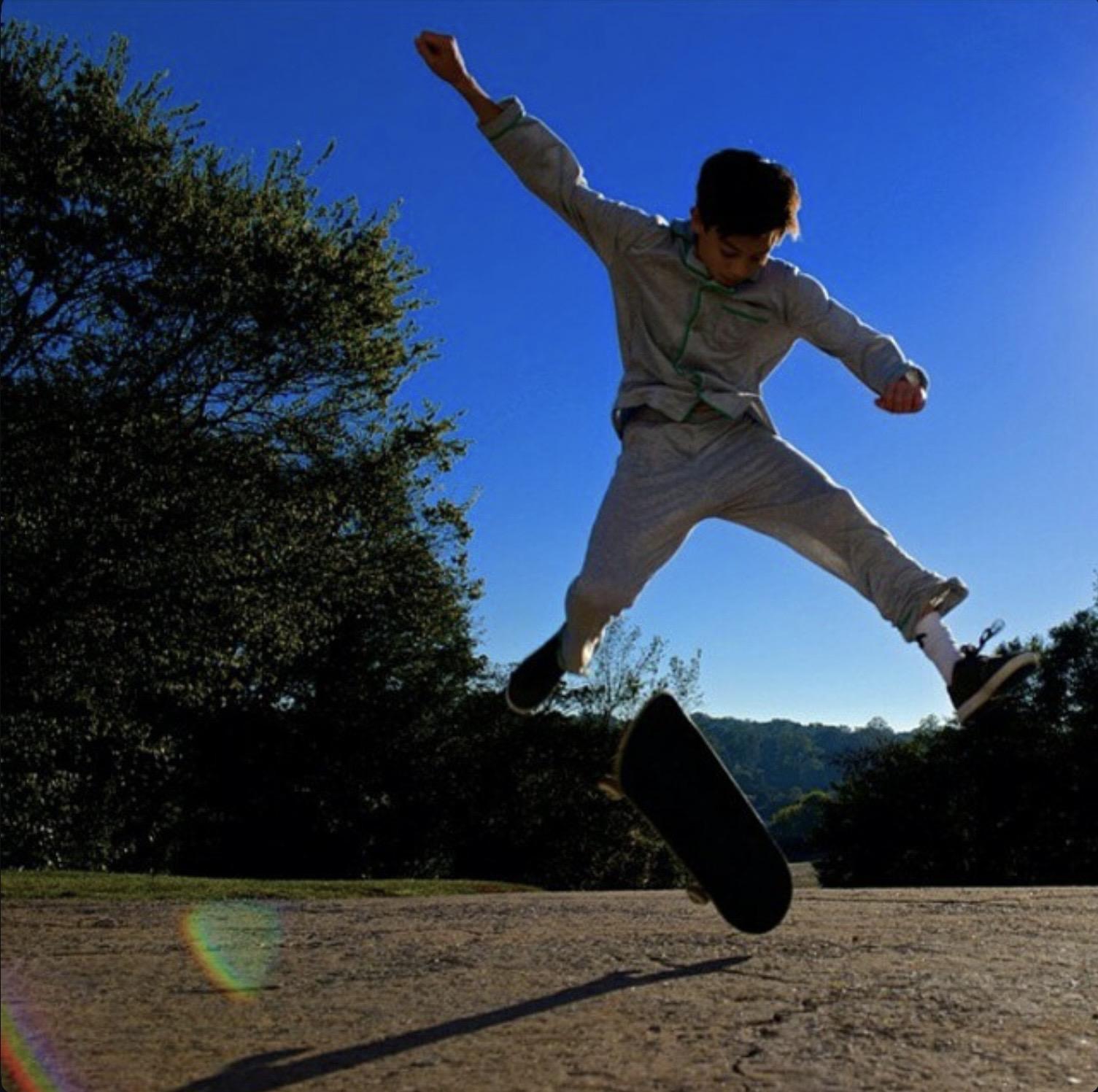 A young teenage boy, wearing light grey pyjamas and black skate shoes, doing a jump from a skateboard.